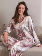 Femme assise en tailleur sur un canapé, portant un pyjama long en satin gris clair avec imprimés floraux roses. Haut boutonné à manches longues avec col passepoilé noir et poche poitrine, assorti à un pantalon fluide. Ensemble doux, élégant et confortable, idéal pour les soirées cocooning avec style.