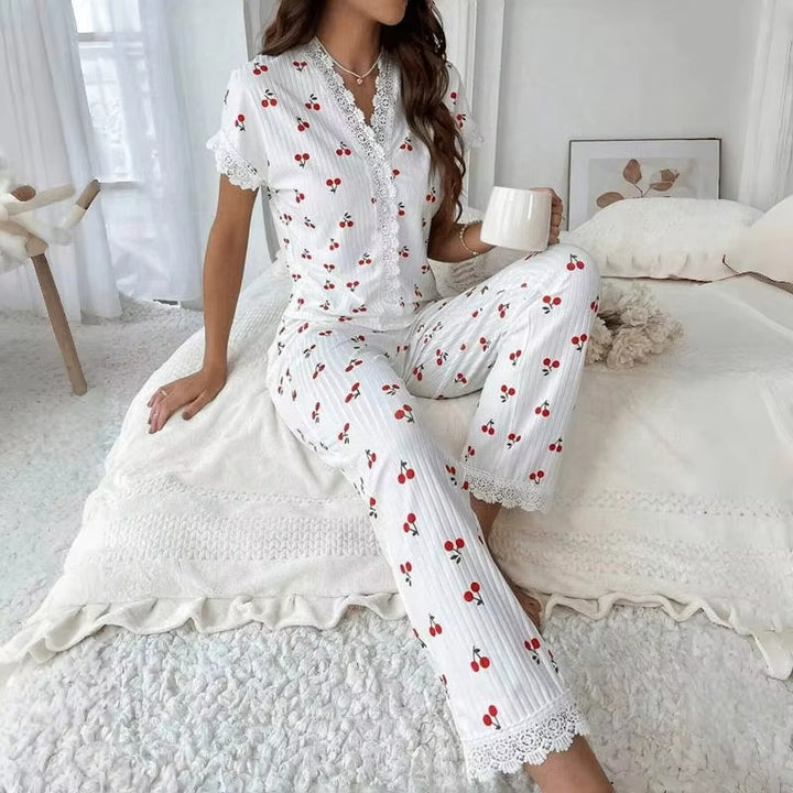 Femme assise sur un lit dans une chambre lumineuse, portant le pyjama Léonie – un ensemble blanc à manches courtes et pantalon long, orné de motifs cerises rouges et de détails en dentelle blanche sur les manches et le col. Pyjama léger et confortable, parfait pour des nuits paisibles.