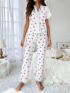 Femme debout dans une chambre lumineuse portant le pyjama "Léonie", un ensemble léger et confortable en tissu blanc à rayures verticales avec motifs de petites cerises rouges. Le haut à manches courtes est orné de dentelle délicate sur le col et les manches, assorti à un pantalon long avec finitions en dentelle aux chevilles. L’ambiance de la pièce est douce et cosy, idéale pour des moments de détente à la maison.