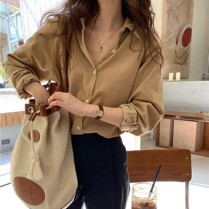 Blouse femme Lana élégante et confortable, coupe fluide à manches longues, coloris beige avec col classique, portée avec un pantalon sombre et un sac en toile pour un look chic et décontracté en intérieur.