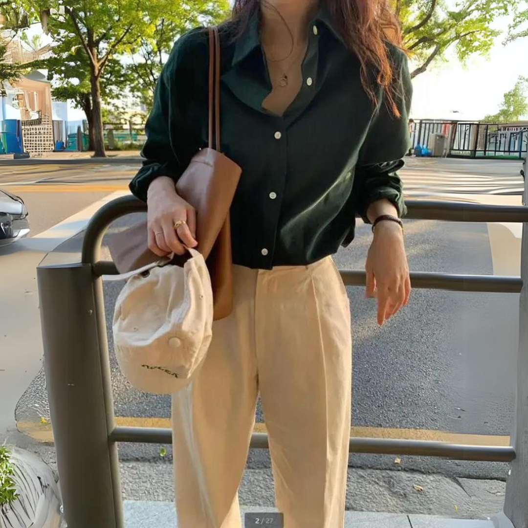 Blouse femme Lana élégante et confortable, coupe fluide à manches longues, coloris vert foncé avec col classique et boutons, portée avec un pantalon beige et un sac en cuir pour un style chic et décontracté en extérieur.