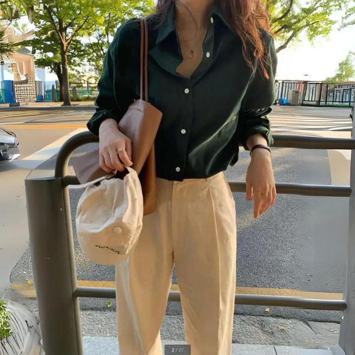 Blouse femme Lana élégante et confortable, coupe fluide à manches longues, coloris vert foncé avec col classique et boutons, portée avec un pantalon beige et un sac en cuir pour un style chic et décontracté en extérieur.