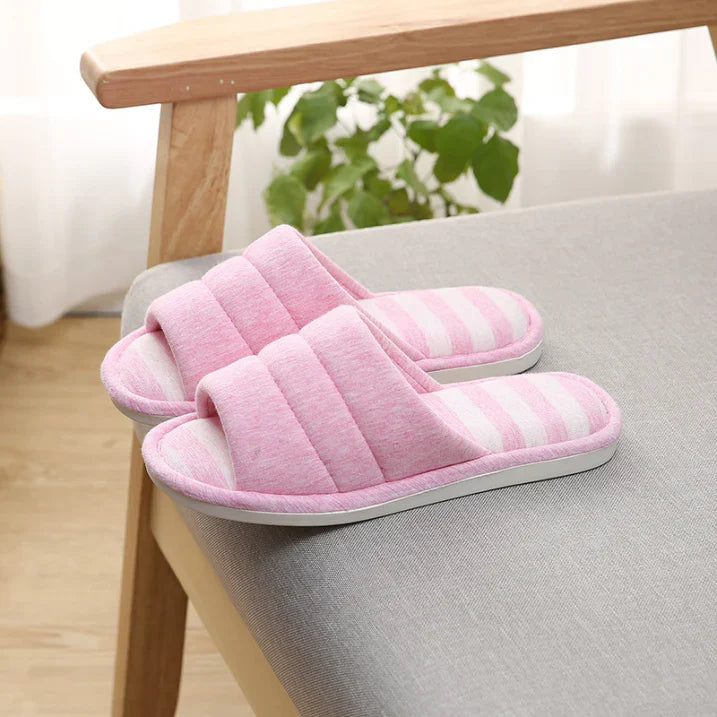 Chaussons femme ouverts rose avec semelle à rayures rose et blanches, posés sur un fauteuil. Texture douce et rembourrée pour un confort cosy, avec semelle antidérapante idéale pour la maison.