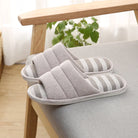 Paire de chaussons femme ouverts gris clair avec semelle à rayures grises et blanches, posés sur un fauteuil. Modèle doux et rembourré pour un confort cosy, avec semelle antidérapante idéale pour la maison.