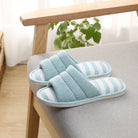 Chaussons femme ouverts bleu clair avec semelle à rayures bleu et blanches, posés sur un fauteuil. Design doux et rembourré pour un confort cosy, avec semelle antidérapante idéale pour la maison.