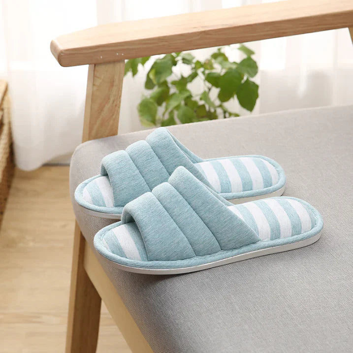 Chaussons femme ouverts bleu clair avec semelle à rayures bleu et blanches, posés sur un fauteuil. Design doux et rembourré pour un confort cosy, avec semelle antidérapante idéale pour la maison.