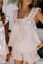 Femme portant un pyjama rose pâle deux pièces, composé d’un débardeur à bretelles larges et volantées, et d’un short assorti à volants. Ensemble satiné, tendance et agréable à porter, idéal pour les journées d'été ou les moments de détente élégants.