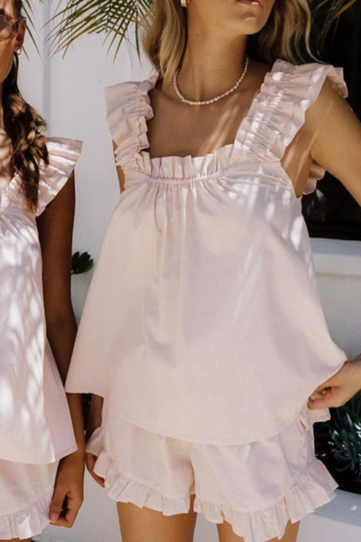 Femme portant un pyjama rose pâle deux pièces, composé d’un débardeur à bretelles larges et volantées, et d’un short assorti à volants. Ensemble satiné, tendance et agréable à porter, idéal pour les journées d&