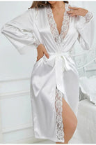 Femme portant un peignoir blanc en satin brillant, décoré de dentelle florale le long du col, des manches et de la fente. Ceinture à nouer à la taille pour un maintien délicat. Modèle chic et raffiné, idéal pour une mariée ou pour une allure élégante à la maison.