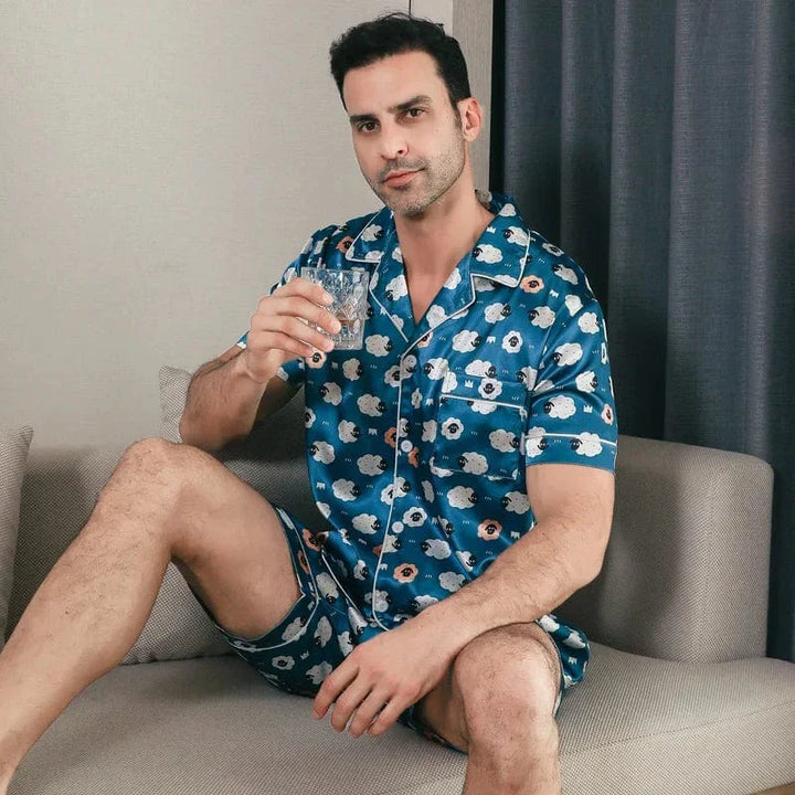 Homme assis portant le pyjama satin Laurent pour homme, ensemble deux pièces avec chemise à manches courtes et short assorti, imprimé bleu à motifs, finition satinée, présenté dans un intérieur moderne.