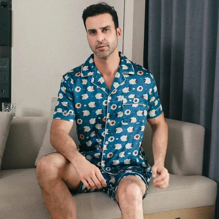 Homme assis portant le pyjama satin Laurent pour homme, ensemble deux pièces avec chemise à manches courtes et short assorti, imprimé bleu à motifs floraux, coupe soignée et finition satinée, présenté dans un intérieur moderne.