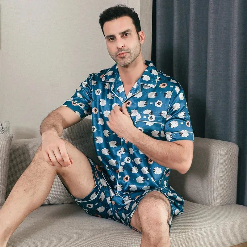 Homme assis portant le pyjama satin Laurent pour homme, ensemble deux pièces avec chemise à manches courtes et short assorti, imprimé bleu à motifs graphiques, finitions contrastées et aspect satiné, présenté dans un intérieur moderne.