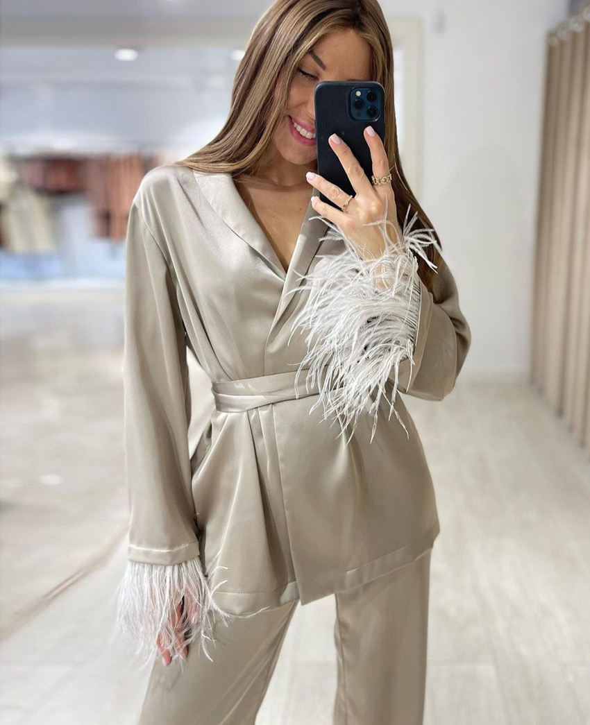 Femme prenant un selfie avec un pyjama satiné beige, orné de plumes blanches aux poignets. Le pyjama est ceinturé à la taille et composé d’un haut à col en V et d’un pantalon fluide, offrant un look à la fois élégant et raffiné, idéal pour des moments de détente ou une soirée chic à la maison.