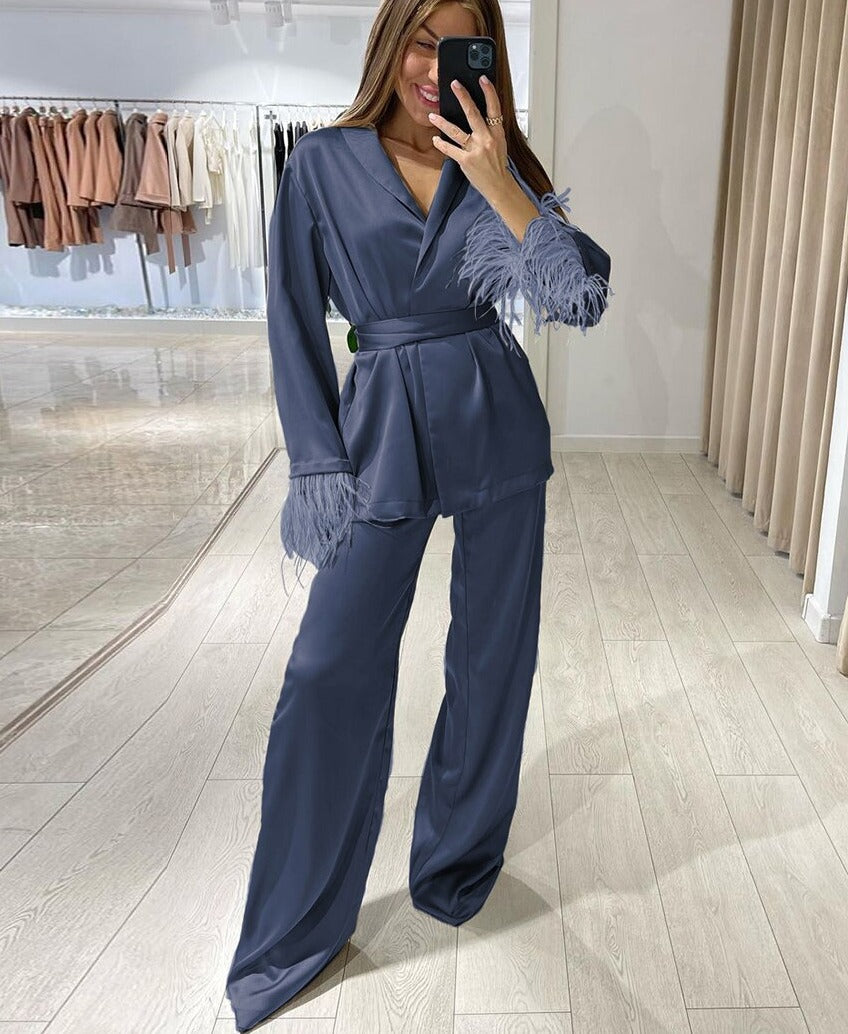 Femme prenant un selfie en pyjama satiné bleu nuit, décoré de plumes ton sur ton aux poignets. Ensemble composé d’un haut croisé à col en V avec ceinture à la taille et d’un pantalon ample et fluide. Un look chic et raffiné, idéal pour une soirée élégante ou une détente luxueuse à la maison.