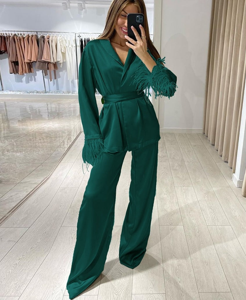 Femme souriante prenant un selfie en pyjama satiné vert émeraude, orné de plumes assorties aux poignets. Ensemble composé d’un haut croisé à col en V, ceinturé à la taille, et d’un pantalon fluide. Un look chic et raffiné, idéal pour une soirée élégante ou un moment de détente luxueux.