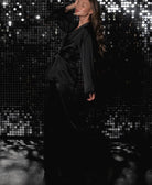 Femme posant avec élégance dans un pyjama satiné noir, ceinturé à la taille et orné de plumes noires aux poignets. Ensemble sophistiqué composé d’un haut croisé à col en V et d’un pantalon fluide, parfait pour une soirée glamour ou une détente chic et raffinée.