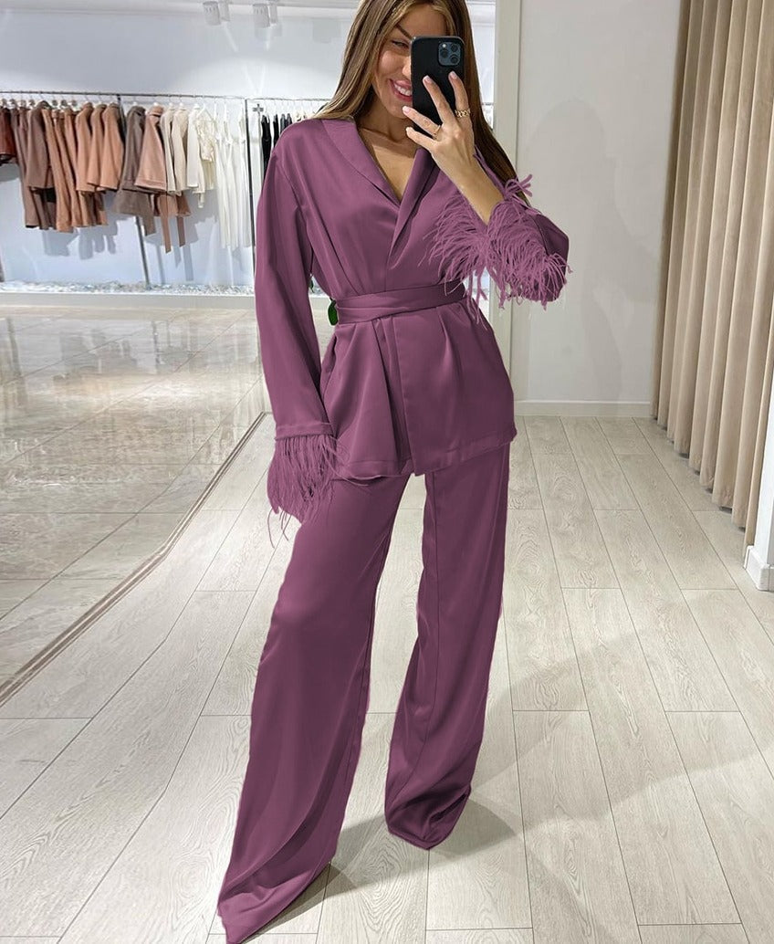 Femme souriante prenant un selfie en pyjama satiné mauve, agrémenté de plumes ton sur ton aux poignets. Ensemble élégant comprenant un haut croisé à col en V, ceinturé à la taille, et un pantalon ample. Idéal pour un look raffiné et chic à la maison ou lors d’une soirée élégante.