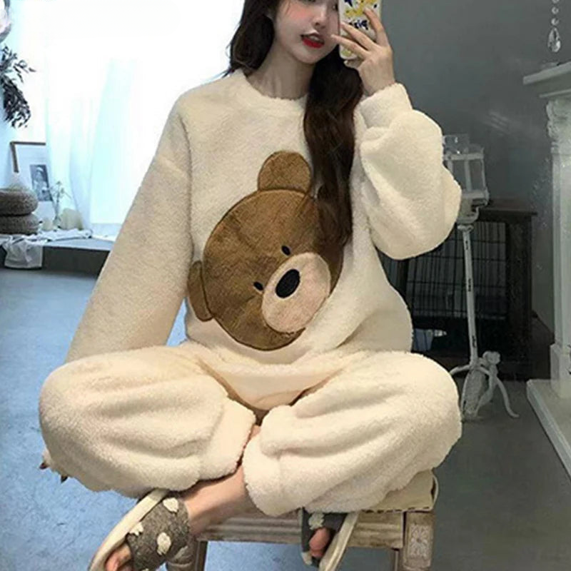 Femme portant un pyjama moelleux blanc cassé avec un grand motif tête d’ours brodé sur le haut à manches longues, assorti d’un pantalon du même tissu. Ensemble doux, chaud et relaxant, parfait pour les moments cocooning à la maison.