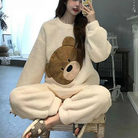 Femme portant un pyjama moelleux blanc cassé avec un grand motif tête d’ours brodé sur le haut à manches longues, assorti d’un pantalon du même tissu. Ensemble doux, chaud et relaxant, parfait pour les moments cocooning à la maison.