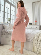 Peignoir femme élégant et confortable Lola  long en tissu doux rose poudré, avec capuche et ceinture à nouer, vue de dos dans une chambre lumineuse.