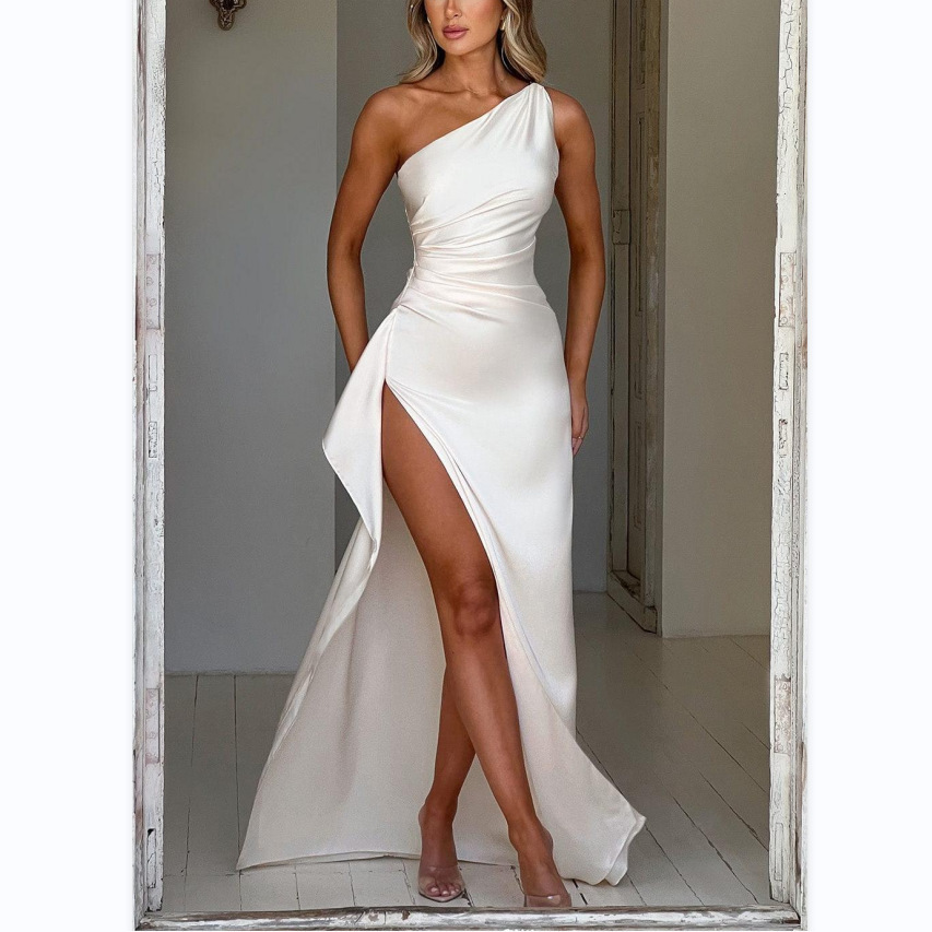 Femme portant la robe satinée Louna en Blanc, avec un design asymétrique à une épaule et une fente latérale audacieuse. Le tissu fluide et brillant crée une silhouette élégante et sophistiquée. Elle est entourée de bougies et de pétales de rose, apportant une touche romantique, parfaite pour une soirée chic. Les chaussures à talons ajoutent un dernier détail glamour.