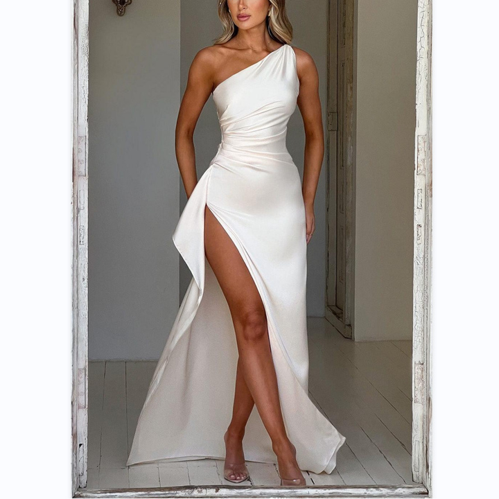 Femme portant la robe satinée Louna en Blanc, avec un design asymétrique à une épaule et une fente latérale audacieuse. Le tissu fluide et brillant crée une silhouette élégante et sophistiquée. Elle est entourée de bougies et de pétales de rose, apportant une touche romantique, parfaite pour une soirée chic. Les chaussures à talons ajoutent un dernier détail glamour.