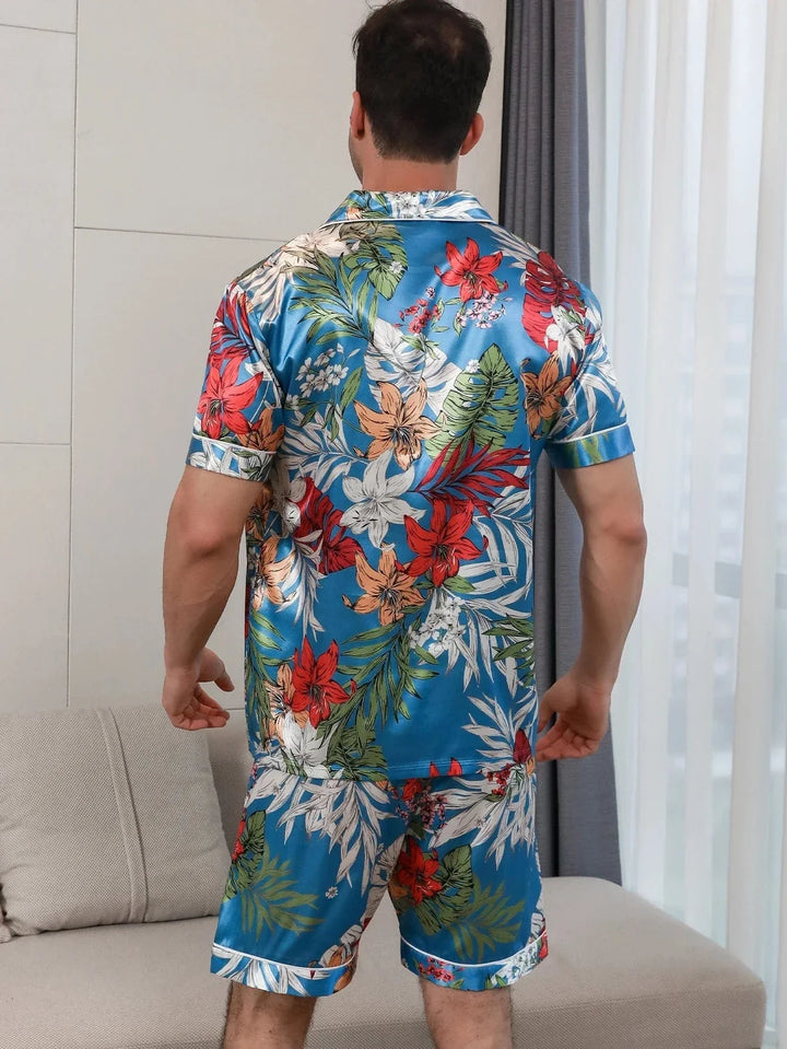 Un homme porte un ensemble de pyjama court en satin à motifs floraux tropicaux, comprenant une chemise à manches courtes et un short assorti. Il est vu de dos, avec des détails floraux colorés sur l&