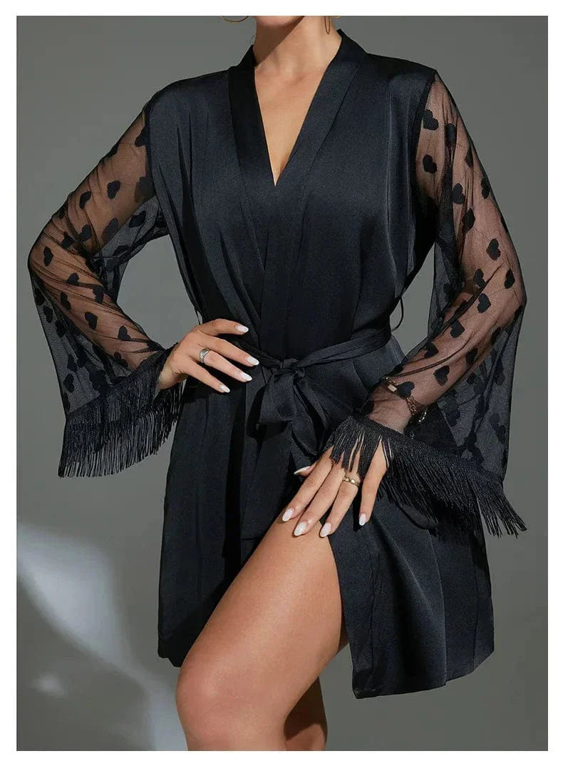 Femme portant le peignoir Lucie noir, avec manches longues en tulle transparent décorées de cœurs et ornées de franges. Ceinture nouée à la taille, tissu satiné et fente sur la jambe. Modèle séduisant et élégant, idéal pour les instants de charme à la maison.