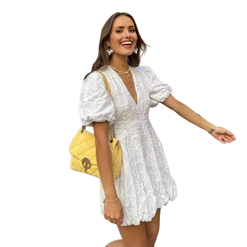 Robe mini satin blanche Lydie portée par une femme souriante en terrasse, coupe fluide et féminine avec manches bouffantes, idéale pour un look élégant et estival.