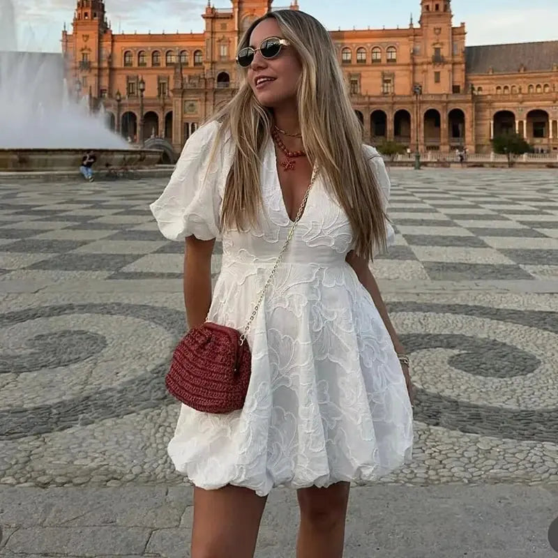 Robe mini satin blanche Lydie portée en extérieur, coupe courte et fluide avec manches bouffantes, style élégant et féminin parfait pour les journées d’été.