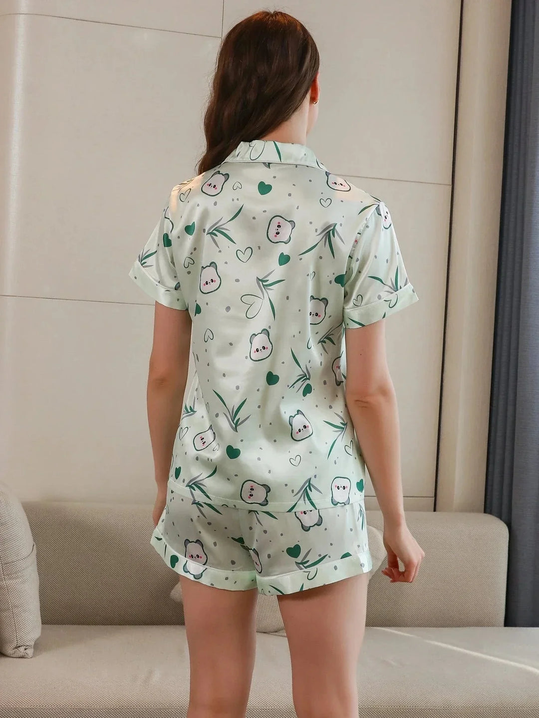 Vue de dos d’une femme portant un pyjama court en satin, composé d’un haut à manches courtes et d’un short assorti, avec imprimés adorables de petits personnages, cœurs et feuillage vert. Ensemble léger, confortable et agréable pour les nuits d&