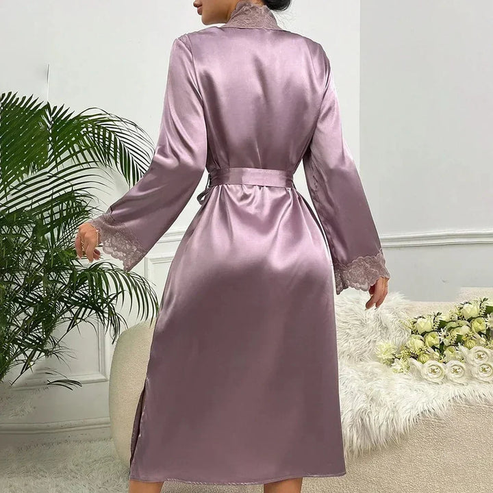 Femme souriante portant le peignoir Mélodie en satin brillant de couleur champagne rosé, à manches longues, avec bordures en dentelle délicate sur les manches et le col. Le peignoir est noué à la taille avec une ceinture assortie, créant une silhouette élégante et féminine. Le tissu soyeux et fluide renforce l’aspect luxueux et confortable du vêtement, parfait pour les moments de détente à la maison.