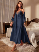 Femme debout dans une chambre, portant une longue nuisette satinée bleu nuit ornée de dentelle beige au col et aux poignets, avec un peignoir assorti. Lingerie élégante, fluide et confortable, idéale pour des soirées cocooning avec style.