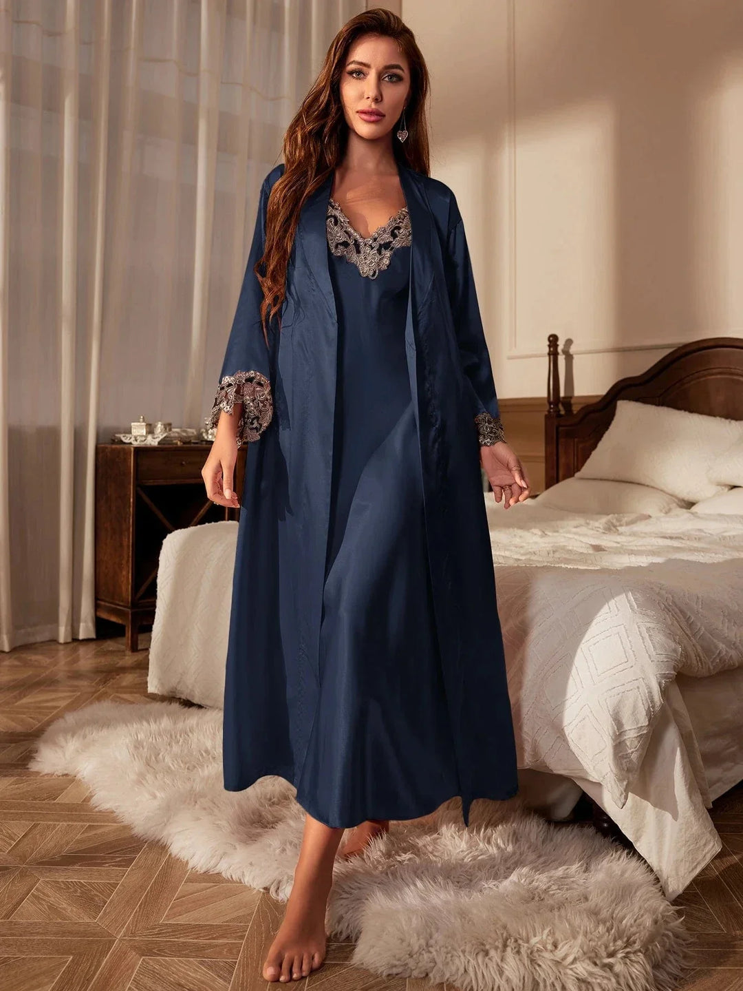 Femme debout dans une chambre, portant une nuisette longue satinée bleu nuit avec empiècements de dentelle beige au col et aux manches, accompagnée d’un peignoir assorti. Ensemble de nuit raffiné, doux et élégant, idéal pour allier confort et glamour à la maison.
