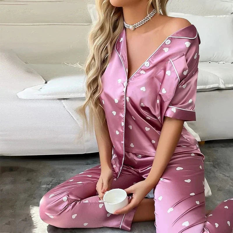 Femme assise tenant une tasse, vêtue du pyjama Pyjama femme Doux, élégant & agréable un ensemble satiné rose décoré de petits cœurs blancs, composé d’une chemise boutonnée et d’un pantalon fluide, idéal pour des moments de détente tout en douceur.