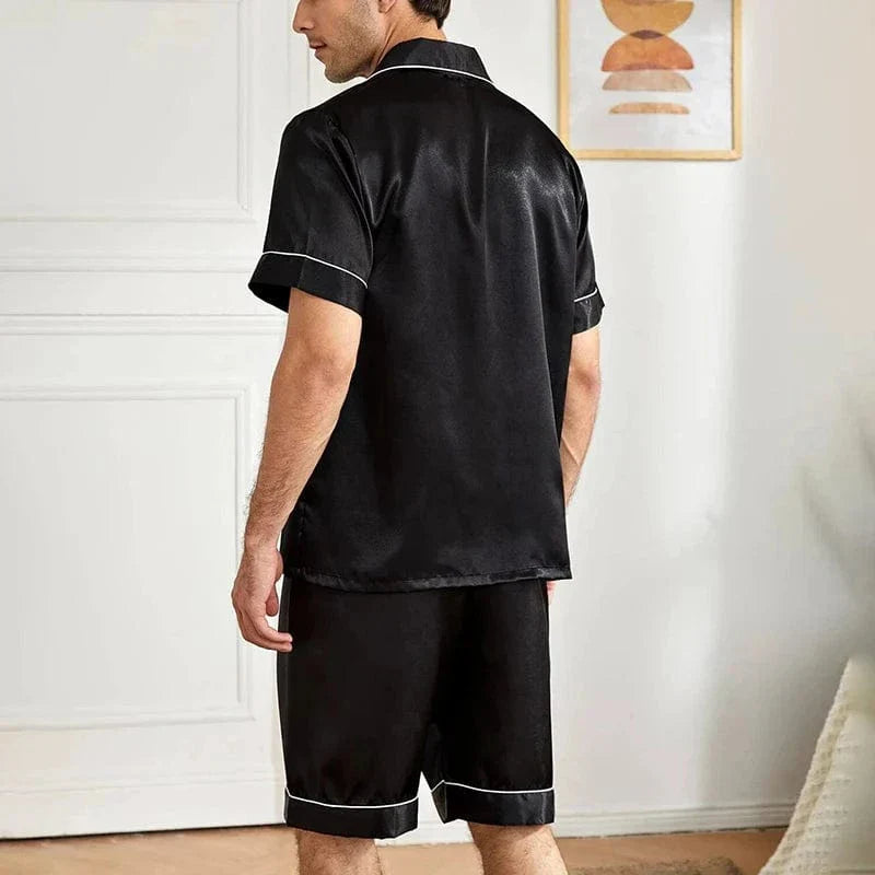 Homme assis au sol portant le pyjama satin pour homme Marcel, ensemble élégant et confortable composé d’une chemise à manches courtes boutonnée et d’un short assorti, en satin noir avec liserés contrastés, offrant une allure raffinée et un confort optimal pour la détente à la maison ou la nuit.