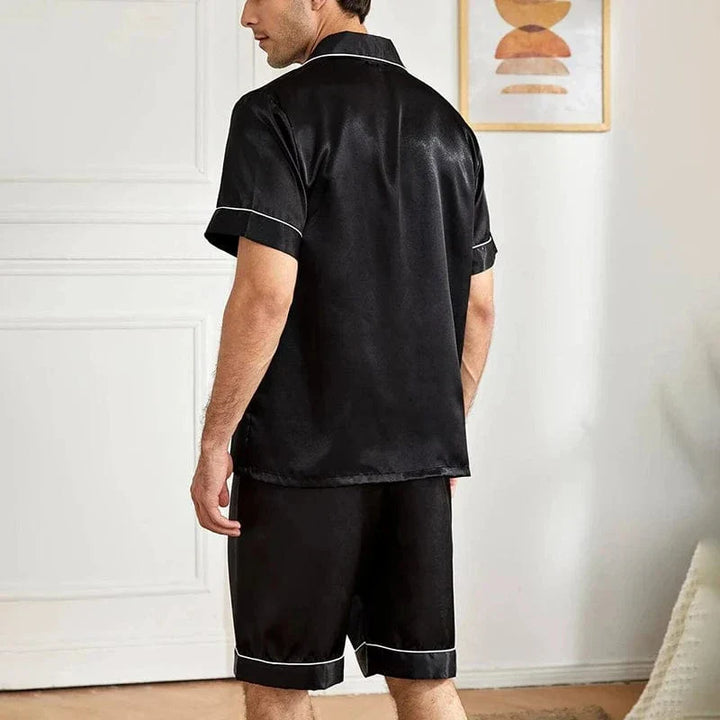 Homme assis au sol portant le pyjama satin pour homme Marcel, ensemble élégant et confortable composé d’une chemise à manches courtes boutonnée et d’un short assorti, en satin noir avec liserés contrastés, offrant une allure raffinée et un confort optimal pour la détente à la maison ou la nuit.