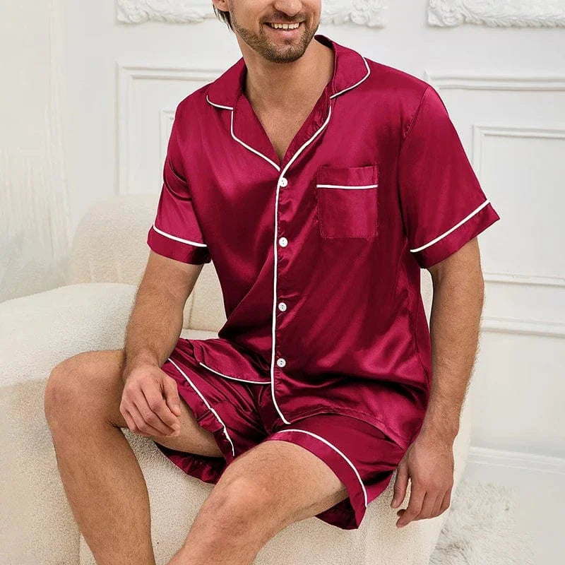 Homme assis sur un fauteuil portant le pyjama satin pour homme Marcel, ensemble élégant et confortable composé d’une chemise à manches courtes boutonnée et d’un short assorti, en satin bordeaux avec liserés contrastés, offrant une allure raffinée et un confort optimal pour la détente à la maison ou la nuit.