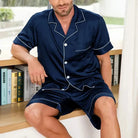 Homme assis sur un meuble portant le pyjama satin pour homme Marcel, ensemble élégant et confortable composé d’une chemise à manches courtes boutonnée et d’un short assorti, en satin bleu marine avec liserés contrastés, offrant une allure raffinée et un confort idéal pour la détente à la maison ou la nuit.