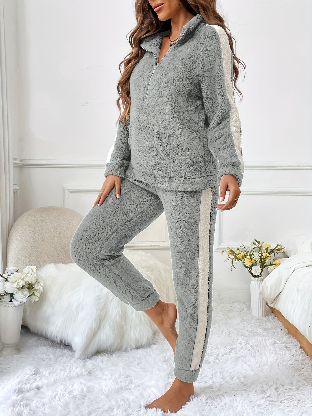 Femme portant un pyjama deux pièces gris clair en matière douce et moelleuse, avec des bandes blanches contrastantes sur les manches et les jambes, posant dans une chambre douillette à la décoration élégante et lumineuse.