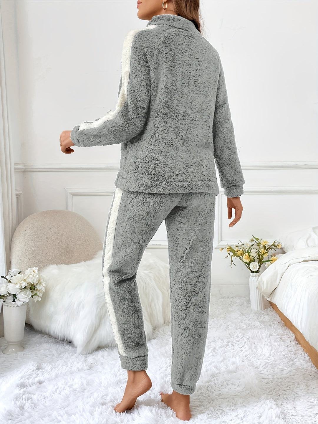 Femme de dos portant un pyjama gris clair deux pièces en tissu pelucheux, avec bandes blanches sur les bras et les jambes, dans une chambre élégante à la décoration cosy avec un lit douillet et un tapis en fausse fourrure.