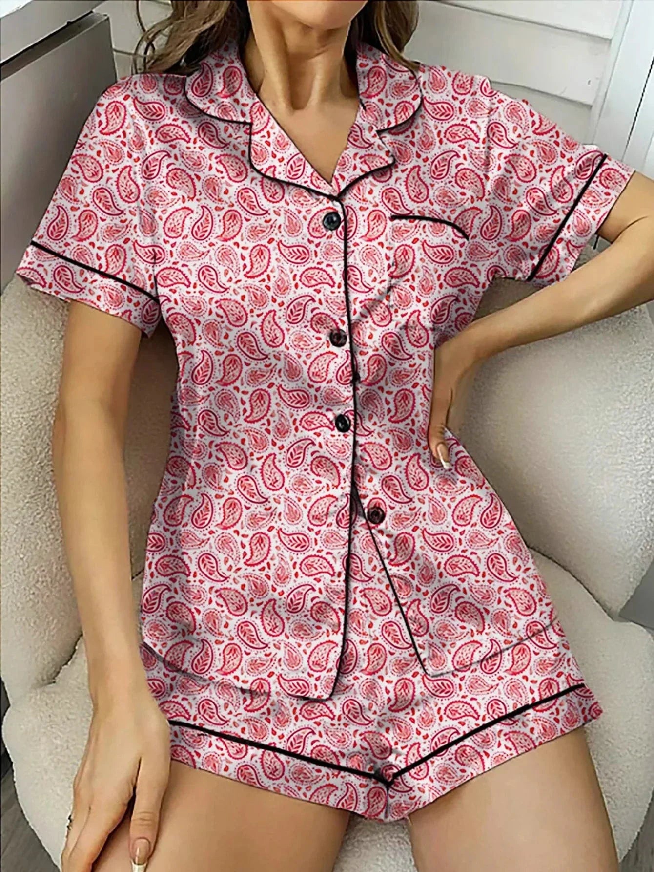 Femme assise portant un pyjama deux pièces à manches courtes, orné de motifs cachemire rouges sur fond clair, avec boutons noirs, col chemise et passepoils contrastants. Ensemble fluide, doux et confortable, idéal pour les nuits estivales et les moments de détente à la maison.