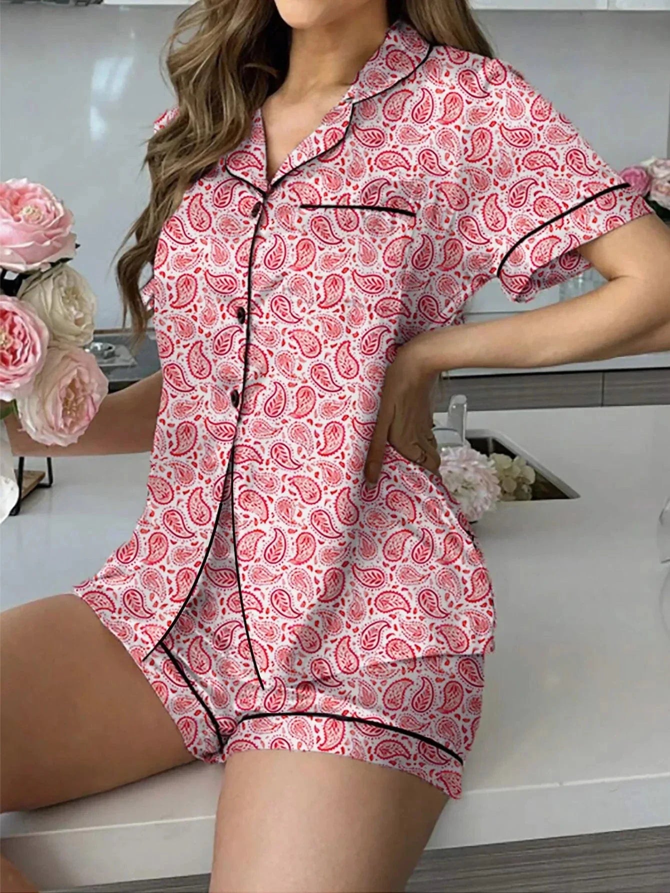 Femme assise portant un pyjama deux pièces imprimé cachemire rouge et blanc, avec haut à manches courtes boutonné, col chemise et passepoils noirs, accompagné d’un short assorti. Ensemble fluide, élégant et confortable, idéal pour des nuits paisibles ou des matinées détendues à la maison.