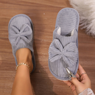 Chaussons femme gris bleuté en velours côtelé avec grand nœud sur le dessus, intérieur doux et chaud. Photo portée, avec un chausson au pied et l’autre tenu à la main, idéal pour rester bien au chaud à la maison.