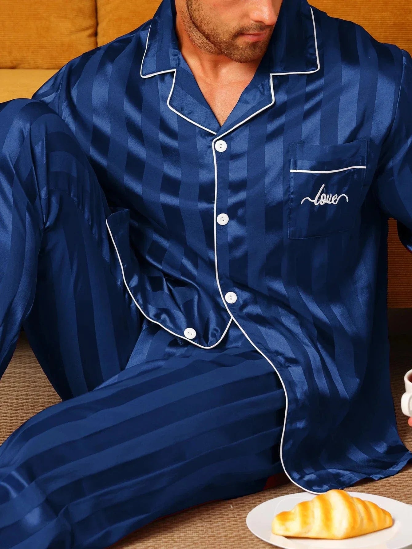 Gros plan d’un homme assis portant le pyjama satin pour homme Maurice, ensemble élégant et confortable en satin bleu marine à fines rayures, avec chemise à manches longues boutonnée, poche poitrine et liserés contrastés, mettant en valeur la texture soyeuse et les finitions raffinées du vêtement.