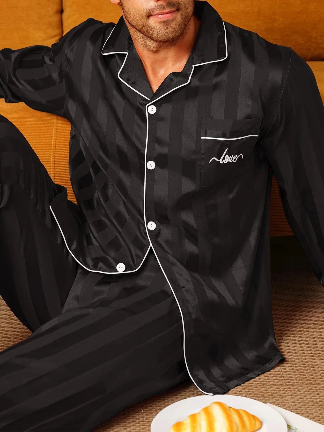 Gros plan d’un homme assis portant le pyjama satin pour homme Maurice, ensemble élégant et confortable en satin noir à fines rayures, avec chemise boutonnée à manches longues, col passepoilé, poche poitrine et finitions contrastées, mettant en avant l’aspect soyeux et raffiné du tissu.