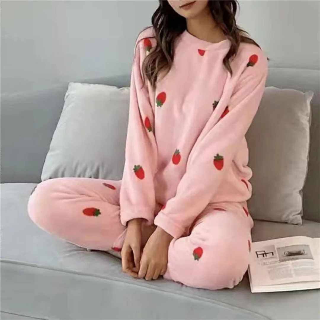 Pyjama femme en peluche rose avec des motifs de fraises rouges, comprenant un haut à manches longues et un pantalon élastiqué. La personne porte le pyjama en position assise sur un canapé gris, avec un livre posé à côté, offrant confort et douceur pour les moments de détente à la maison.