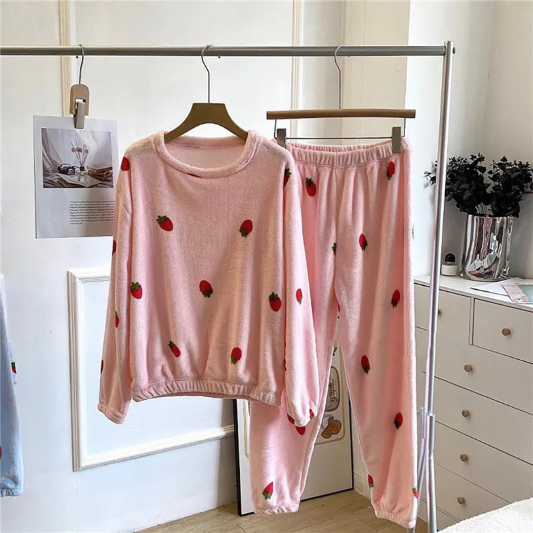 Pyjama femme en peluche rose avec des motifs de fraises rouges, comprenant un sweat à manches longues et un pantalon élastiqué assorti, suspendus sur un porte-vêtements, offrant confort et douceur pour des moments de détente.