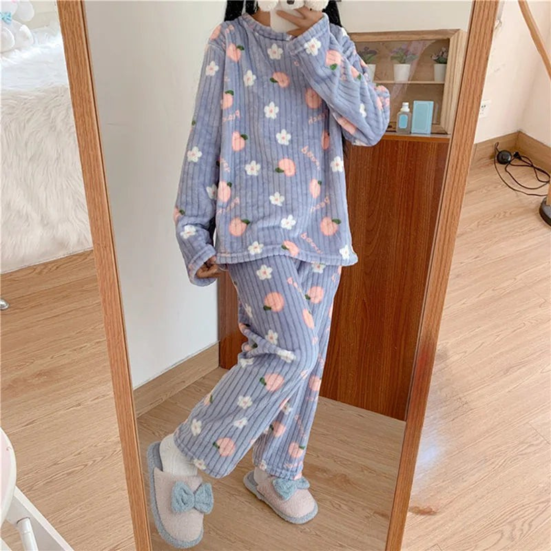 Pyjama femme en peluche douce de couleur bleue, décoré de motifs de fleurs blanches et de pêches roses. La personne porte le pyjama en se tenant devant un miroir, avec des chaussons doux et confortables à nœud, offrant un look cosy et tendance pour se détendre à la maison.