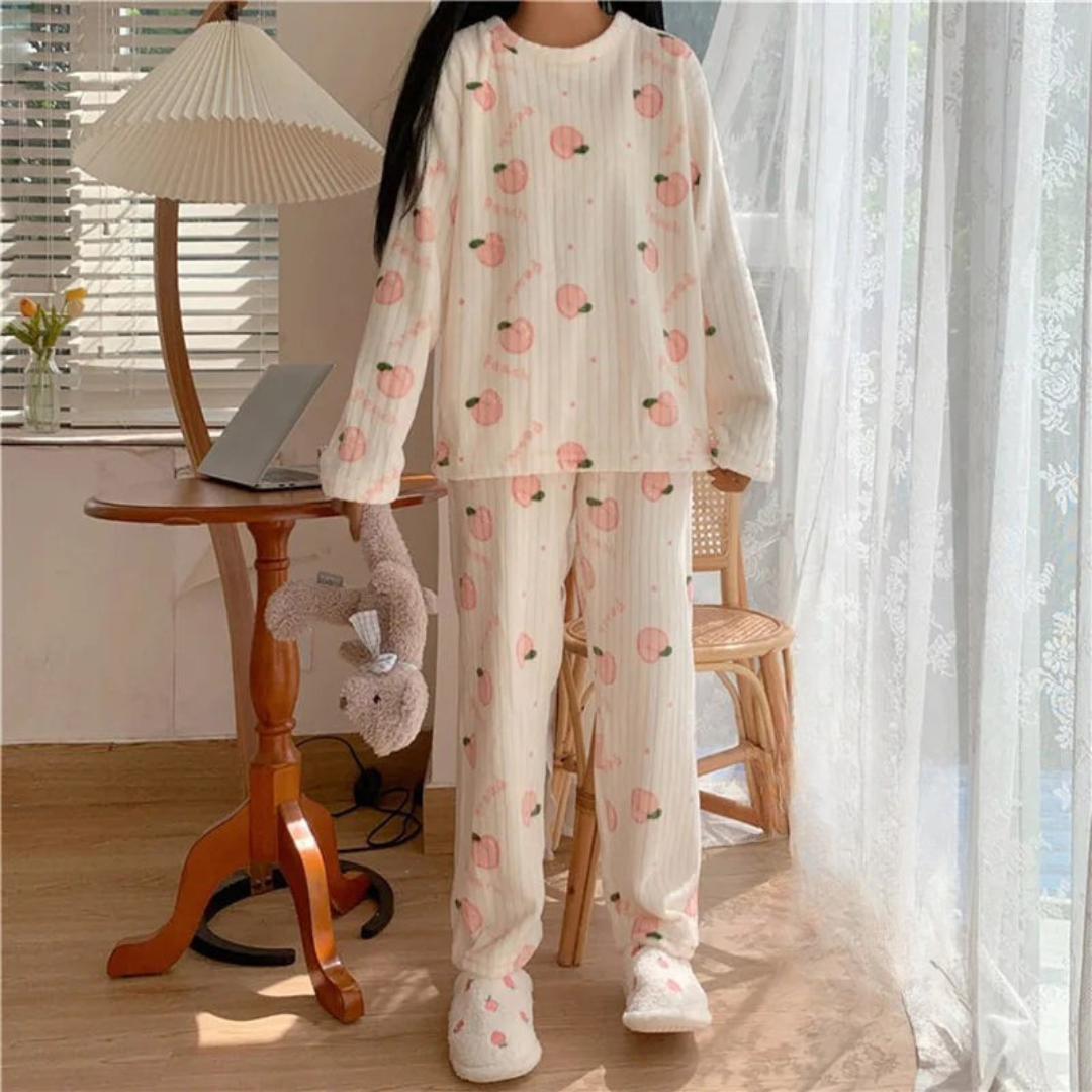 Pyjama femme en peluche douce de couleur blanche avec des motifs de pêches roses. La personne porte le pyjama et se tient debout près d'une fenêtre, avec des chaussons blancs confortables et un doudou en peluche. Le look est cosy et parfait pour se détendre à la maison.